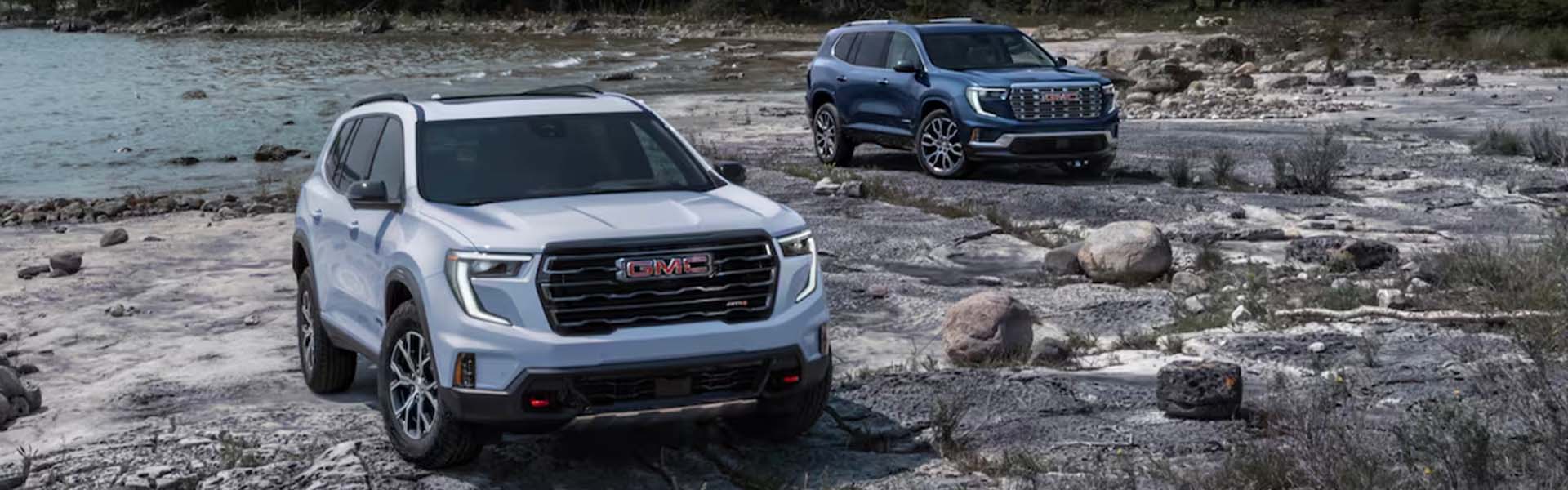 2025 GMC Acadia midsize SUVs shown off-road near a rocky lakeshore, featuring a rugged exterior design.