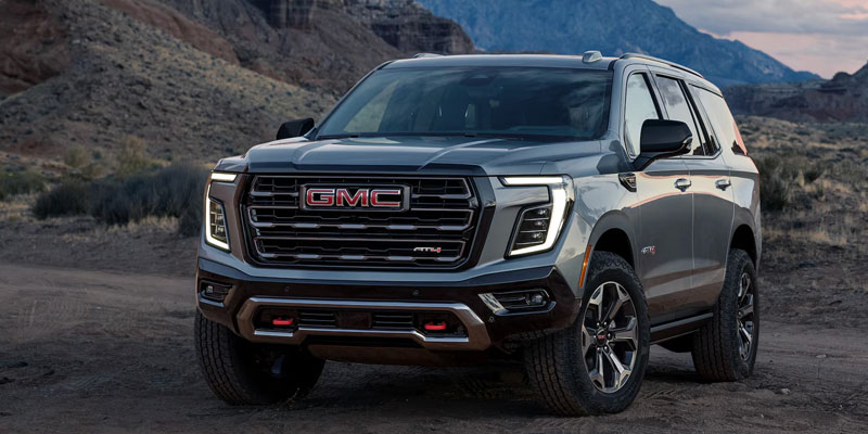 A sleek, black GMC Yukon AT4 is parked on rugged terrain with mountains in the background at dusk. The vehicle's bold grille and modern design exude strength.