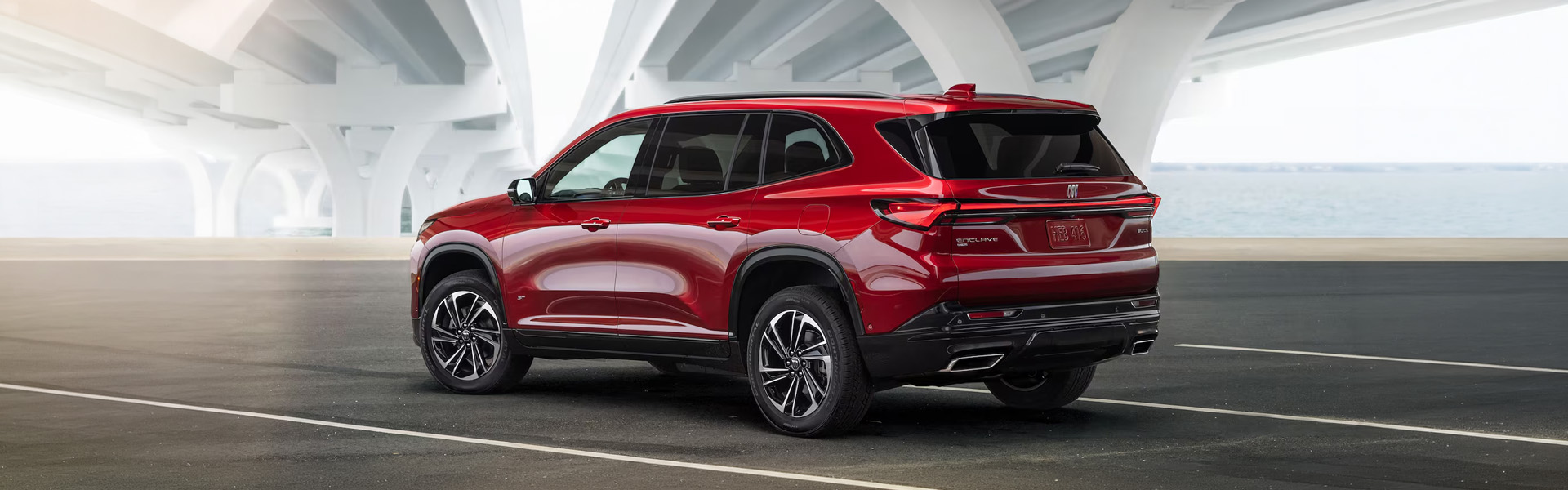 A 2026 Buick Enclave in a deep red color, parked under a modern overpass with water in the background.