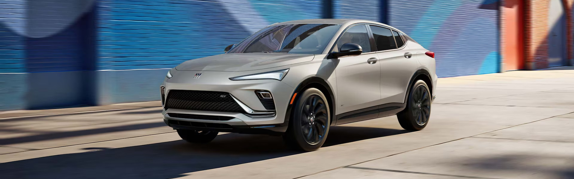 2026 Buick Envista ST compact crossover in white, driving along a city street with a colorful mural backdrop, featuring a sporty grille, black wheels, and sleek LED headlights.