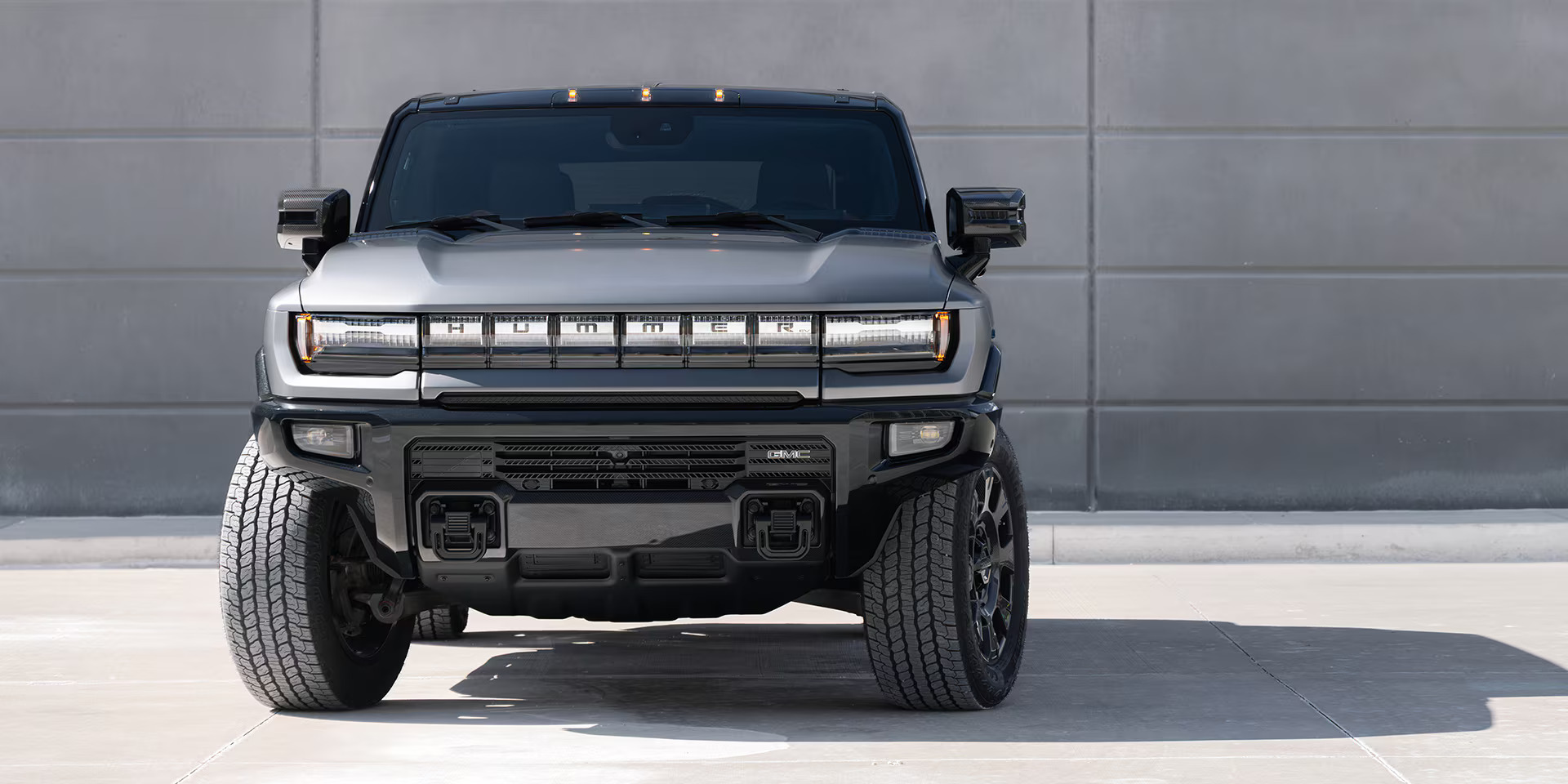 Front view of the 2026 GMC Hummer EV SUV in silver, showcasing its bold grille, signature LED lighting, rugged off-road stance, and wide tires parked against a modern industrial backdrop.